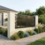 Metalowy panel ogrodzeniowy w ciemnym kolorze z ażurowym wzorem liści zamontowany między jasnymi słupkami przy nowoczesnym domu.