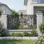 Metalowy panel ogrodzeniowy w czarnym kolorze z klasycznym ornamentalnym wzorem zamontowany między jasnymi słupkami przy eleganckiej posesji.