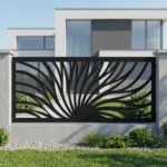 Metalowy panel ogrodzeniowy w czarnym kolorze z dynamicznym ażurowym wzorem promieni zamontowany między jasnymi słupkami przy nowoczesnym domu.