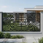Metalowy panel ogrodzeniowy w czarnym kolorze z dynamicznym ażurowym wzorem falistych linii zamontowany między jasnymi słupkami przy nowoczesnym domu.