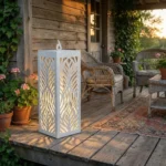 Metalowy lampion ogrodowy Cień Natury w kolorze białym z ażurowym roślinnym wzorem ustawiony na drewnianym tarasie w ogrodzie